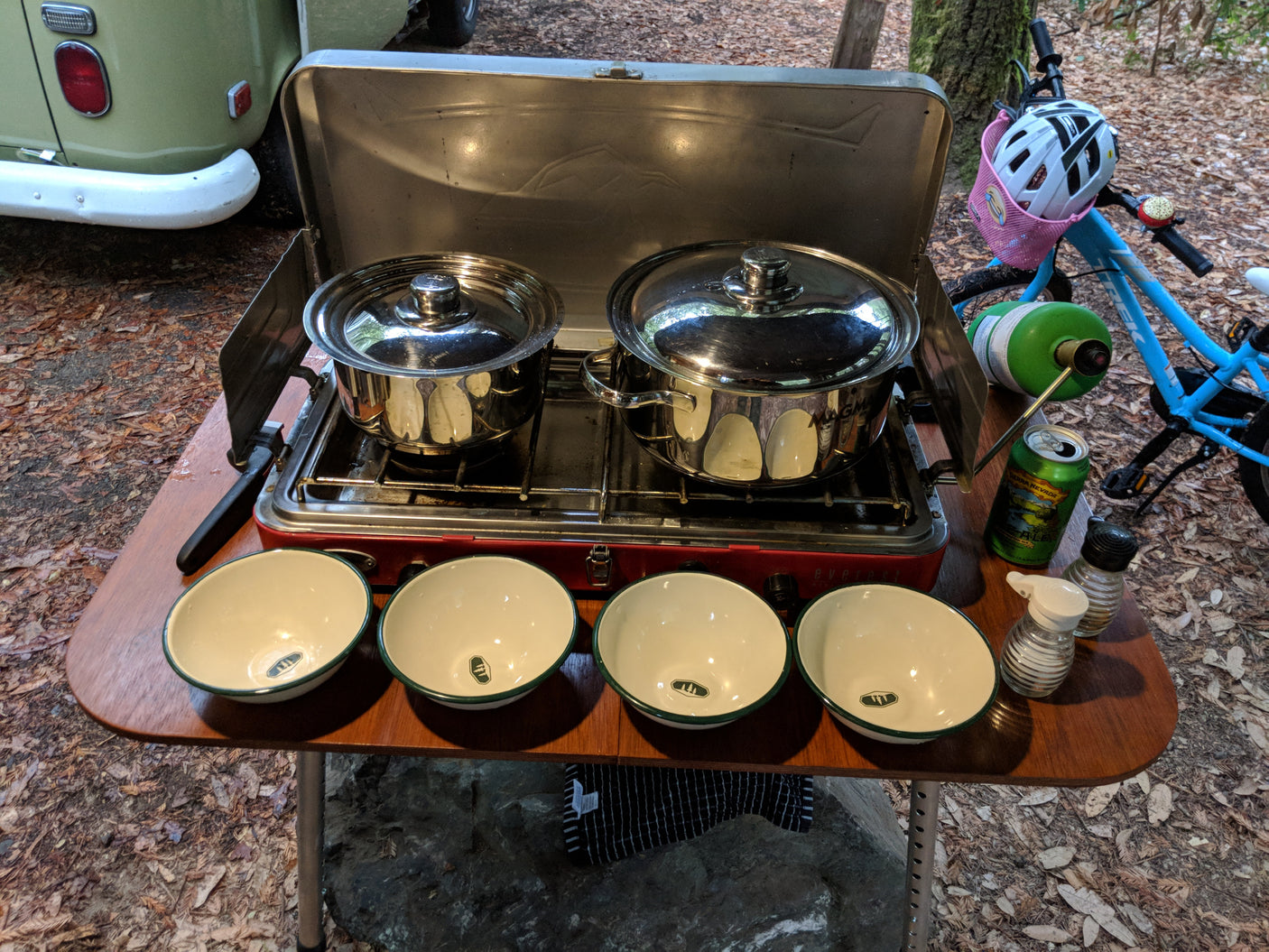 RV rental outdoor cooking: A camp stove with pots, bowls, beer, and spices on a table, plus an RV and bike.