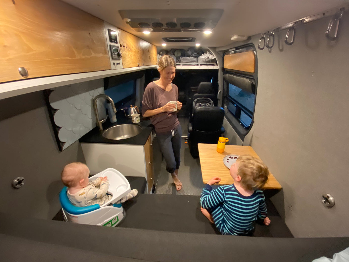 Inside a comfortable RV rental: Mom with coffee, a baby in a high chair, and a toddler eating at a small table.