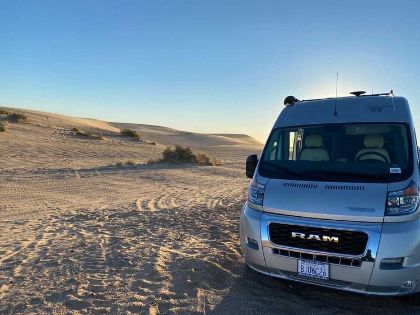 Exploring desert dunes in a silver RAM RV rental. Van parked on sand with tire tracks and distant dunes.