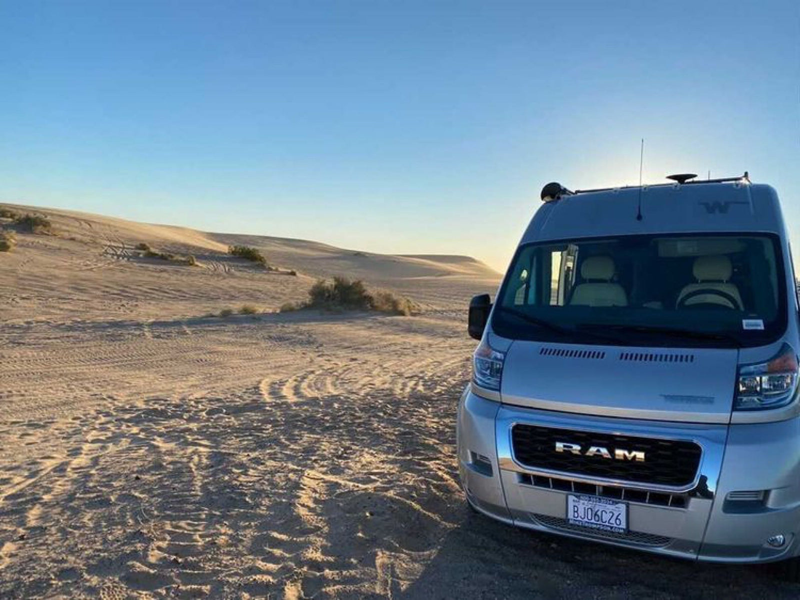 Exploring desert dunes in a silver RAM RV rental. Van parked on sand with tire tracks and distant dunes.