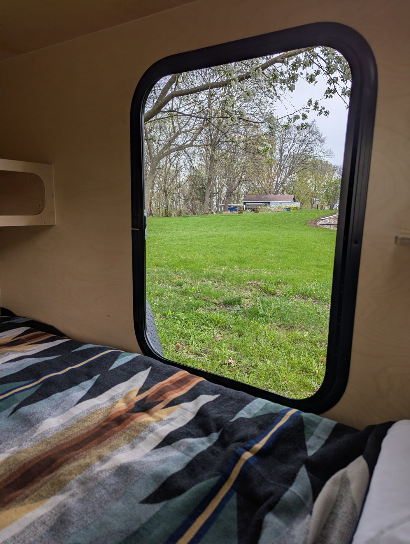 Enjoy nature from this RV rental! Cozy bed with a patterned blanket, looking out a window to a green yard and blooming trees.