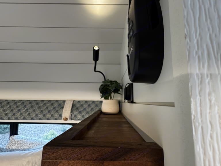 Cozy RV rental interior featuring a bed, small plant, reading light, and control panel on white shiplap walls.