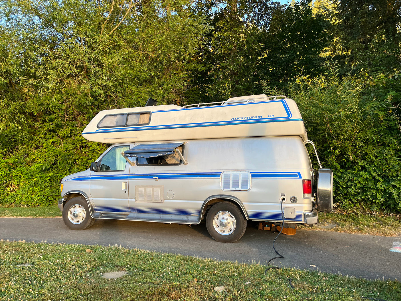 Silver Airstream 190 camper van, potentially an RV rental, parked on a paved path by a lush treeline with power hookup.