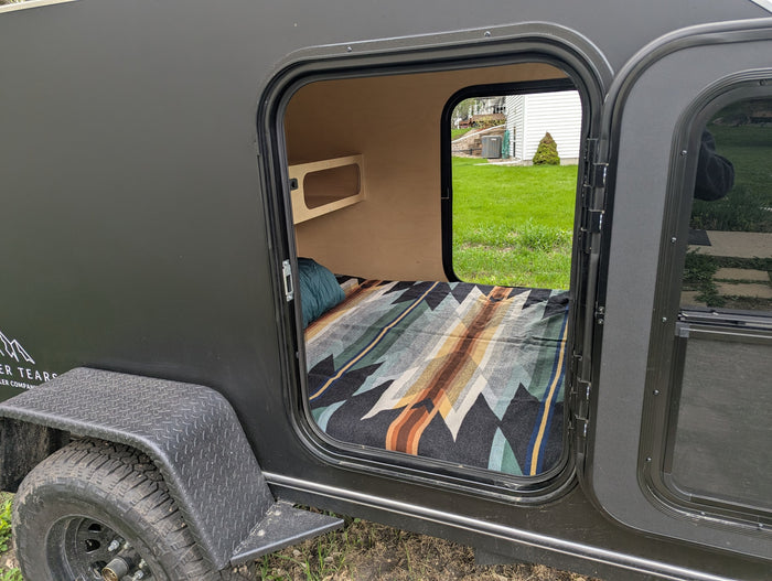 View into a dark grey teardrop camper's open door, showing a cozy bed and shelf. Great for an rv rental adventure.