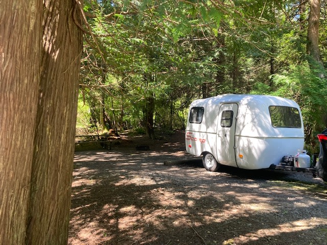 A small white RV rental is parked on a shaded dirt campsite