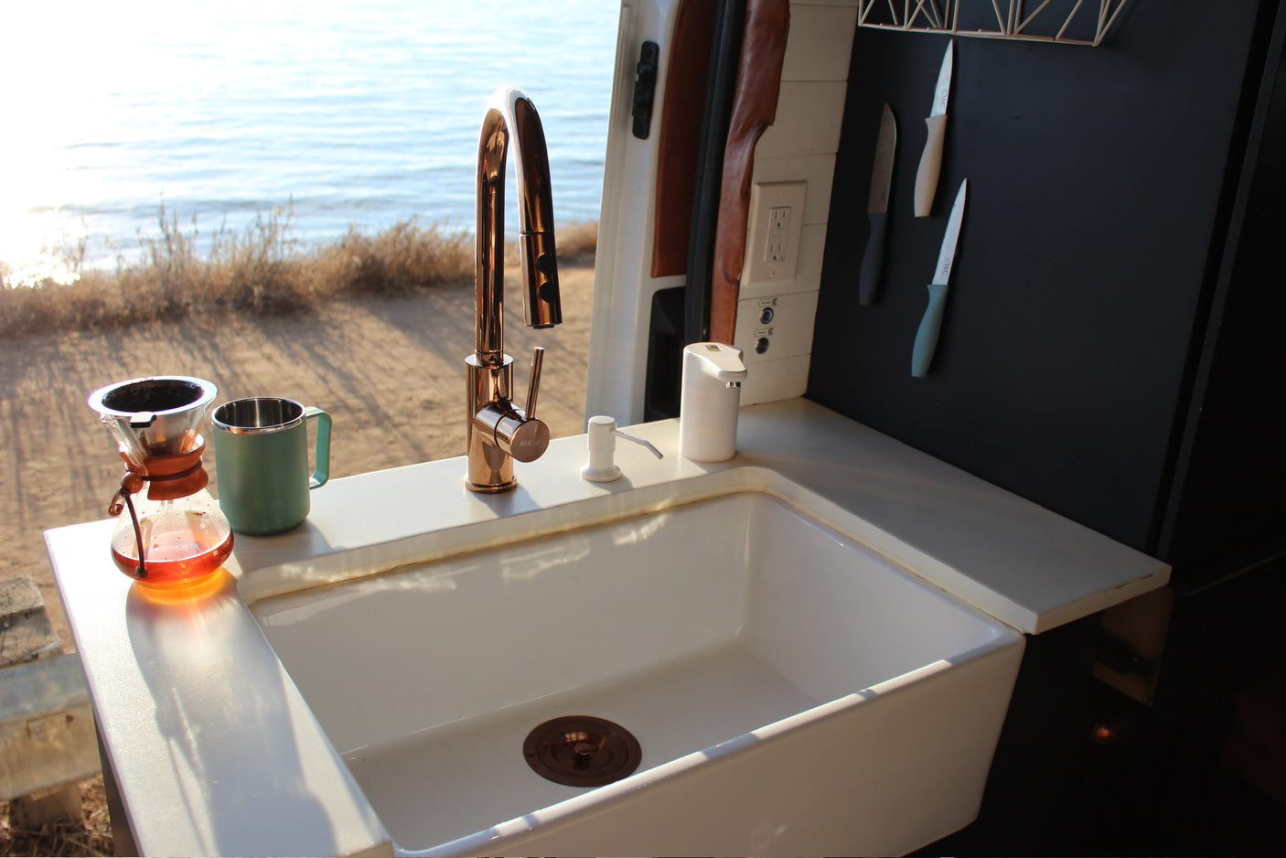 Interior of an RV rental kitchen with a large white sink, rose gold faucet, coffee setup, and ocean view.