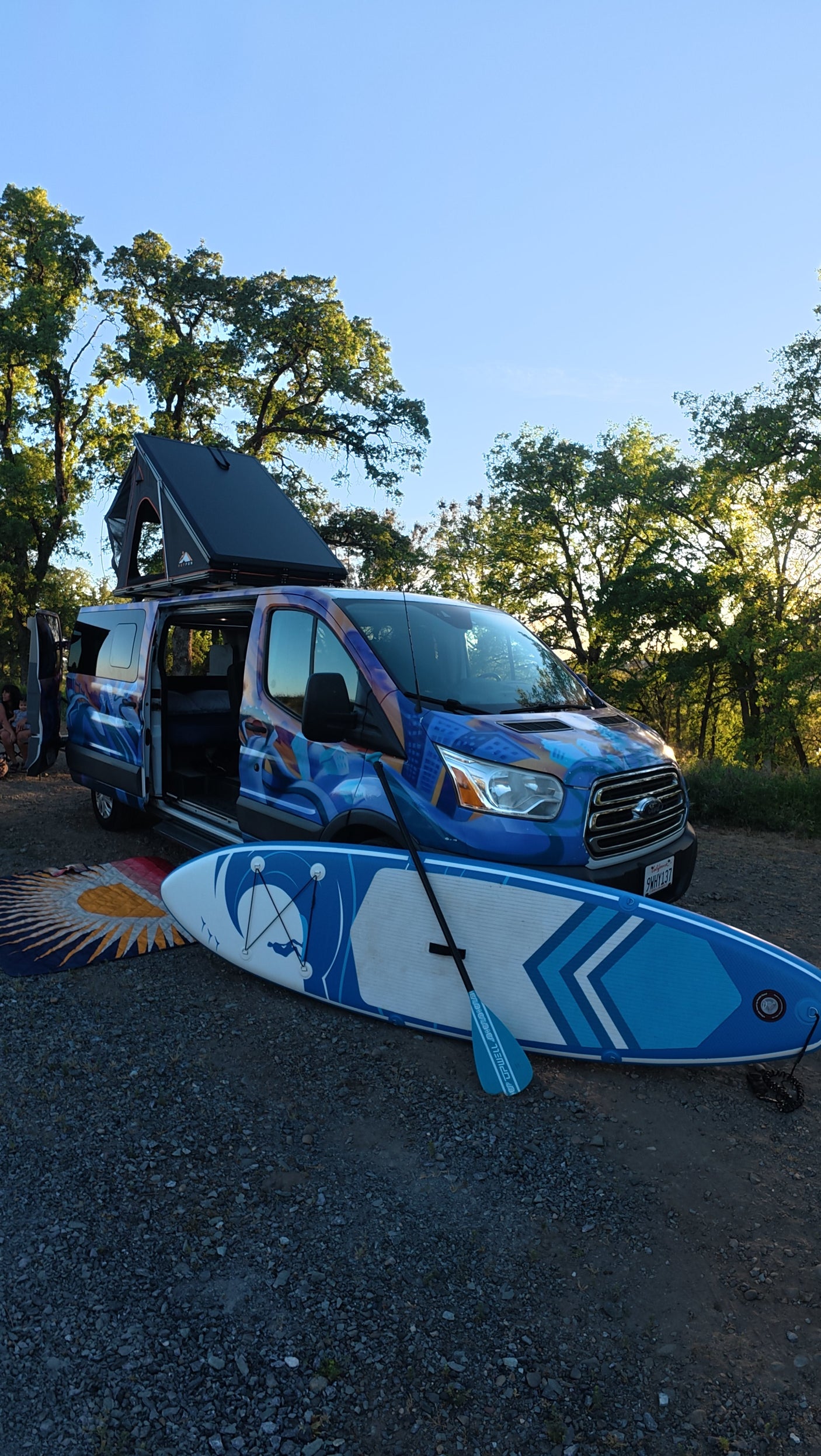 Blue/purple custom RV rental camper van with pop-top tent & paddleboard on gravel. Perfect for outdoor adventures!