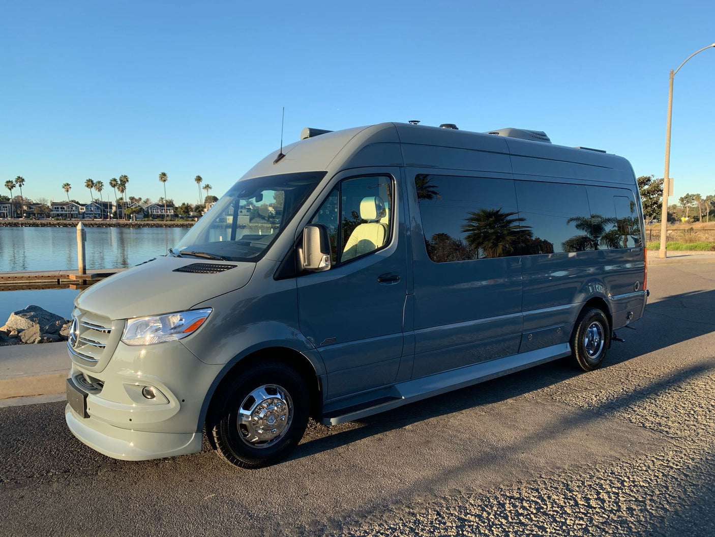 Light grey Mercedes-Benz Sprinter rv rental camper van parked by a sunny waterfront with palm trees and houses.