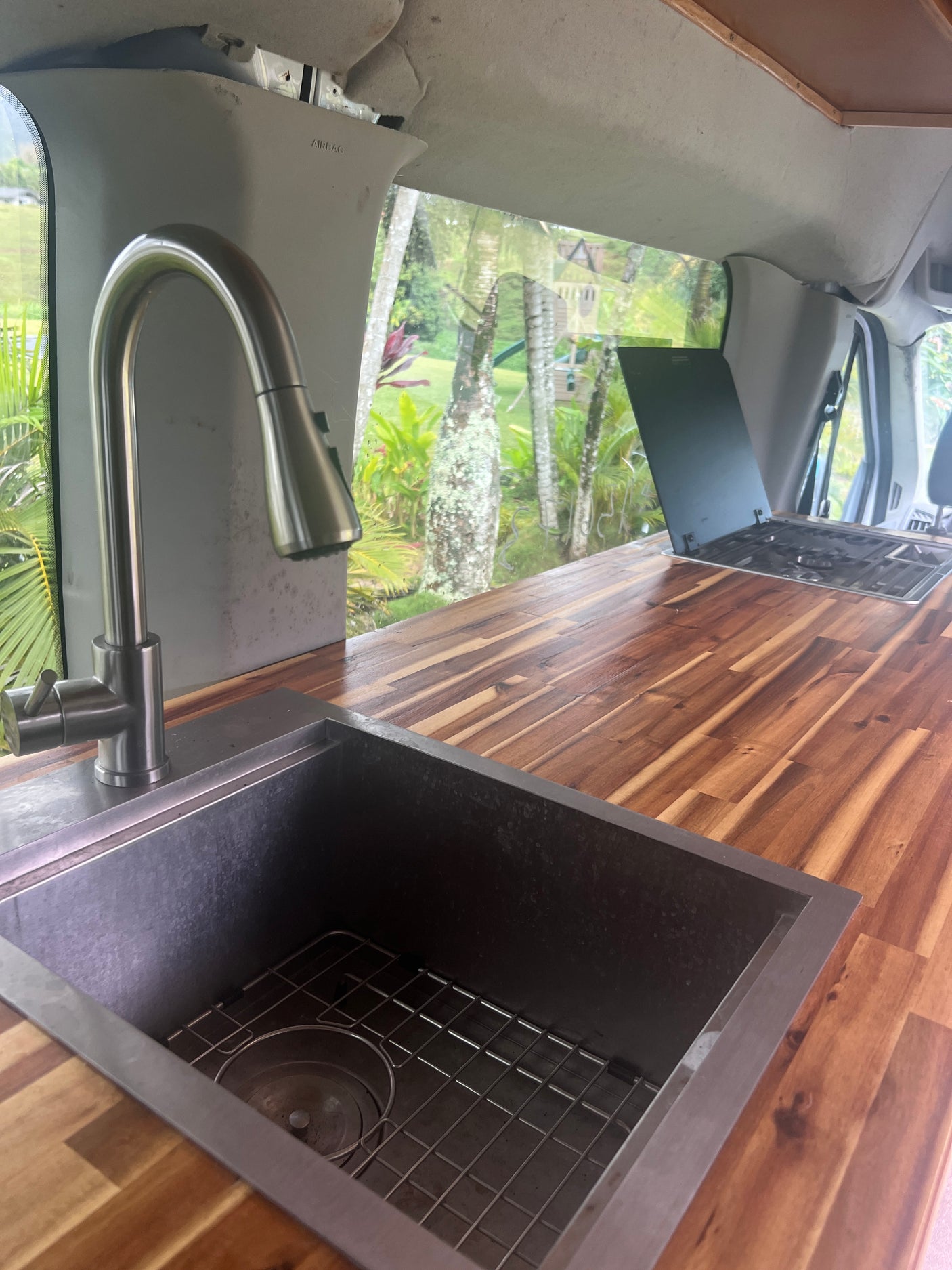 Inside an RV rental: a modern van kitchen with a stainless steel sink, wood countertop, and scenic tropical view.