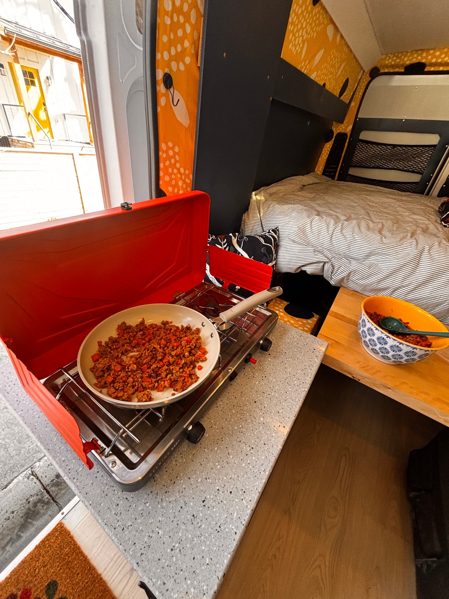 Preparing a hot meal inside an RV rental. Skillet of ground meat and veggies on stove, bed in background of the camper.