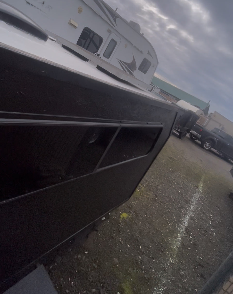 Side view of a black vessel/trailer parked, with a white RV rental behind it on a gravel lot under a cloudy sky.