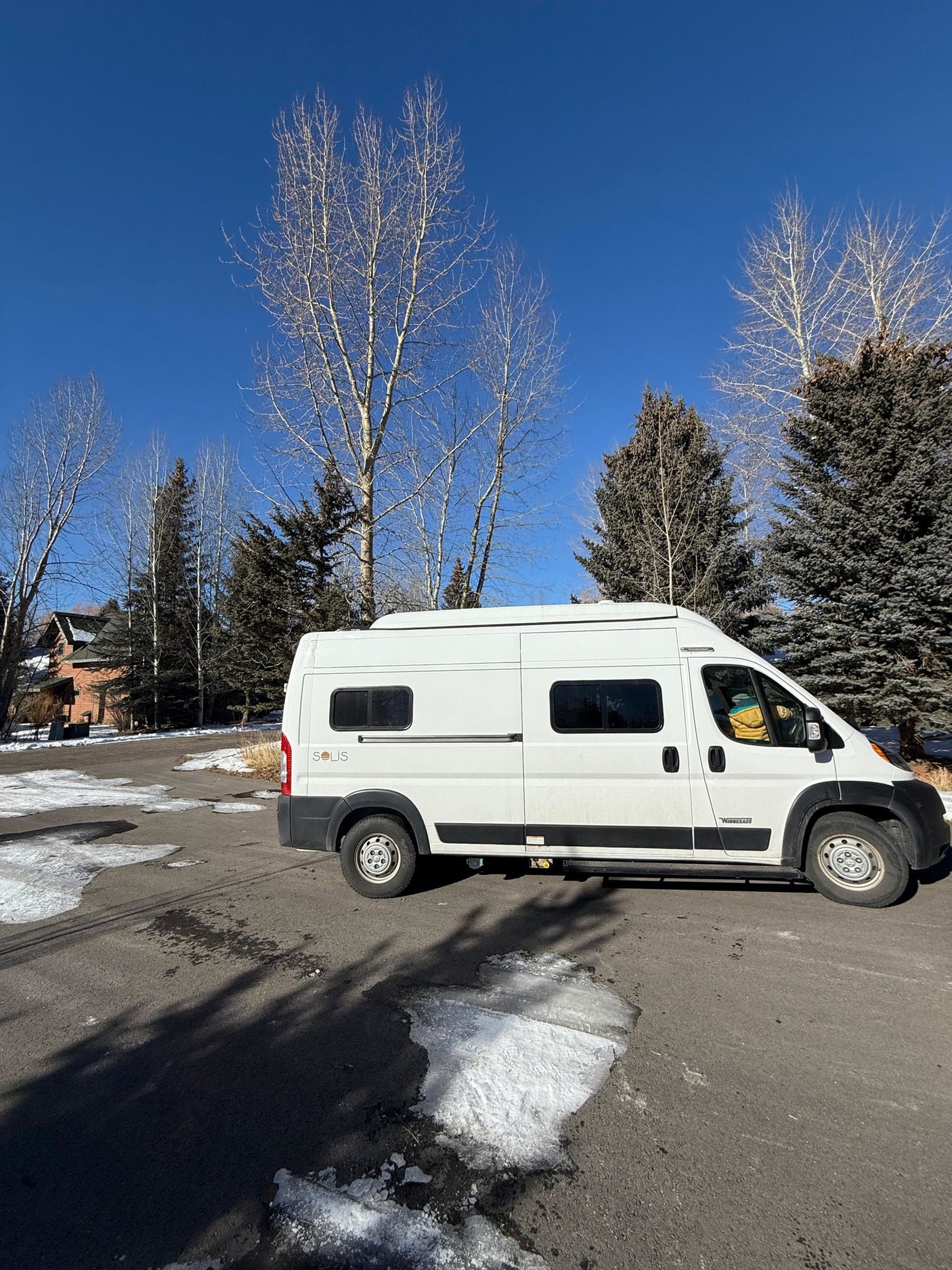 A white Winnebago Solis rv rental van is parked on an icy lot with bare trees and evergreens under a bright blue sky.