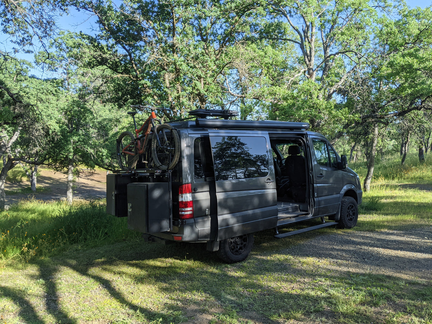 Explore the outdoors with this gray camper van RV rental, equipped with a mountain bike, parked under trees.