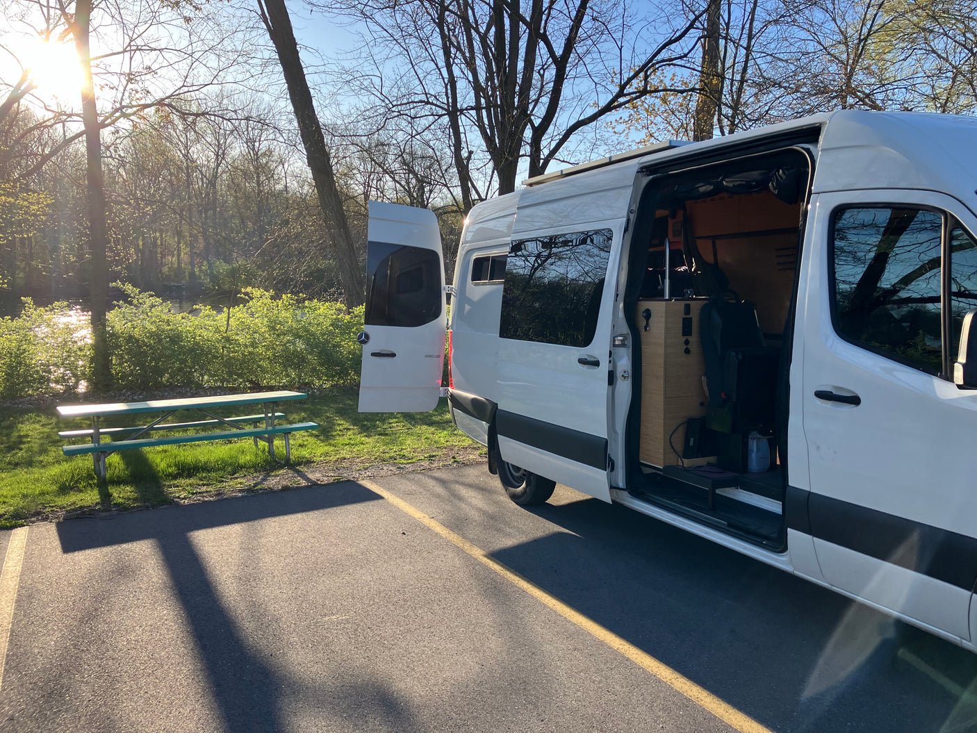 A white camper van, perfect for your next RV rental, is parked near a tranquil river and picnic table under sunlight.