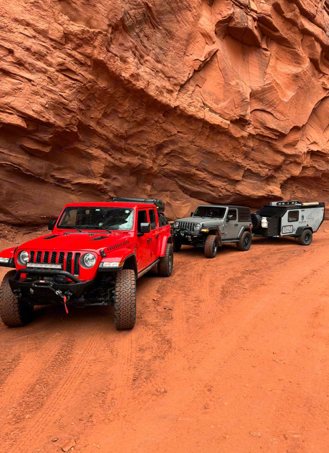 Two Jeeps lead a rugged RV rental camper trailer on a red dirt trail, surrounded by towering rock formations, ideal for adventure.