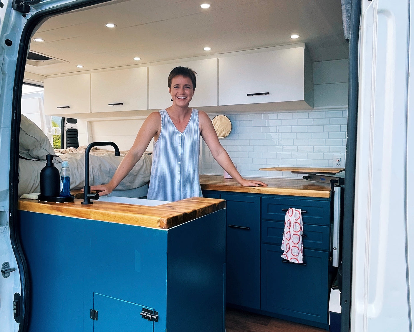 Smiling woman in a spacious RV rental kitchen featuring blue base cabinets, wood countertops, and white tiled backsplash.