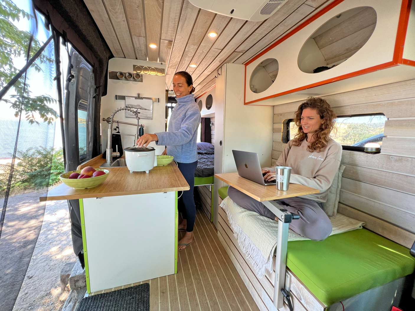 Two women in an RV rental: one cooking at the kitchen, the other working on a laptop in a cozy, bright camper van.