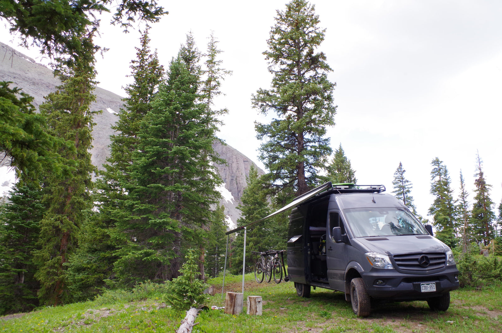 An RV rental, a grey Mercedes Sprinter van, is set up with an awning & bikes in a mountain forest campsite.