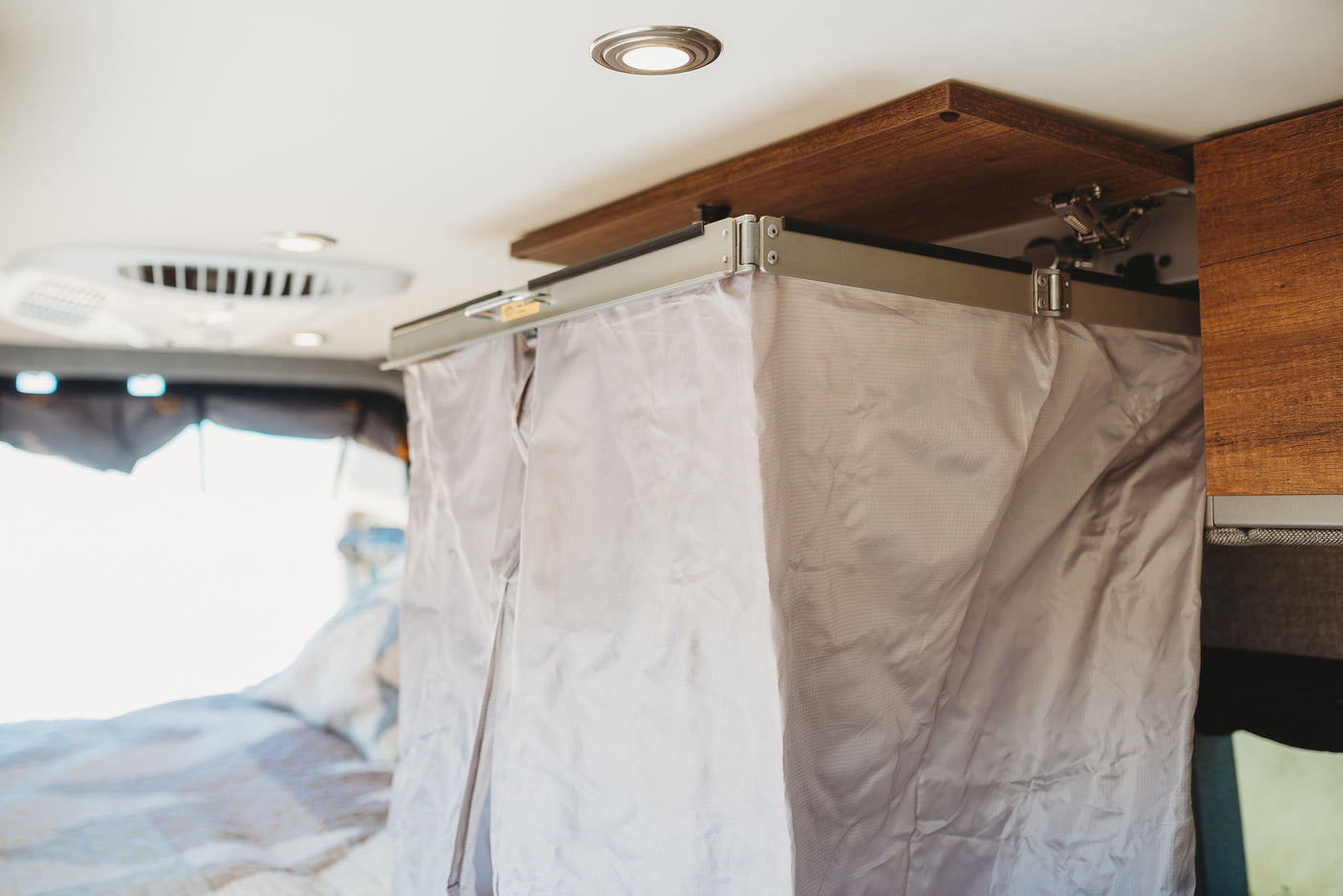 Inside an RV rental: a light-gray privacy curtain hangs from a ceiling track, with a wooden storage shelf above and recessed lights.