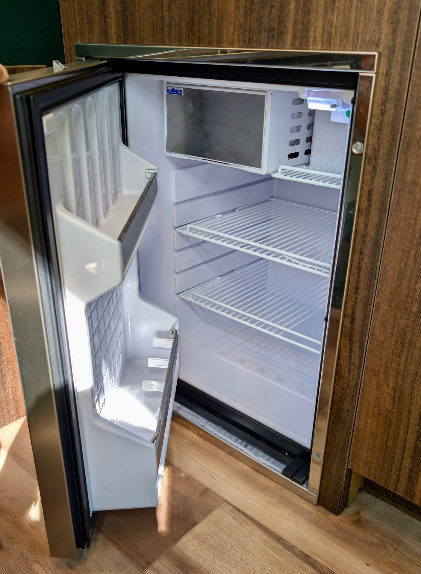 An open, empty mini-fridge inside an RV rental. Features include wire shelves, a small freezer, and integrated design within wooden cabinetry.