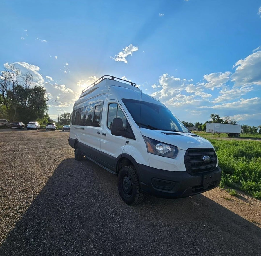 Ready for your next adventure! This white Ford Transit rv rental camper van sits on a gravel lot under golden sunrays.|RV rental fun! A smiling person peeks from a white pop-top campervan. Perfect for camping and outdoor travel adventures.|Modern RV rental interior: tiled shower wall