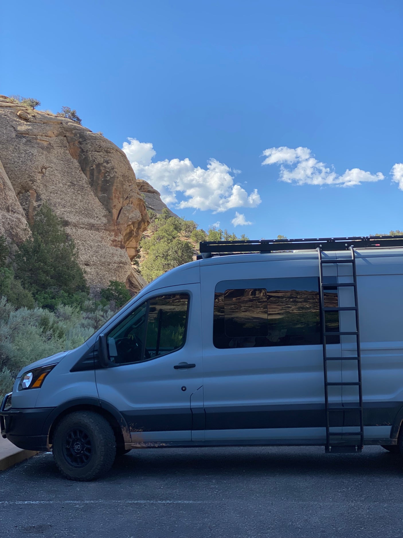 A grey camper van (an RV rental) with a ladder and roof rack is parked near a massive rocky cliff under a blue, cloudy sky.