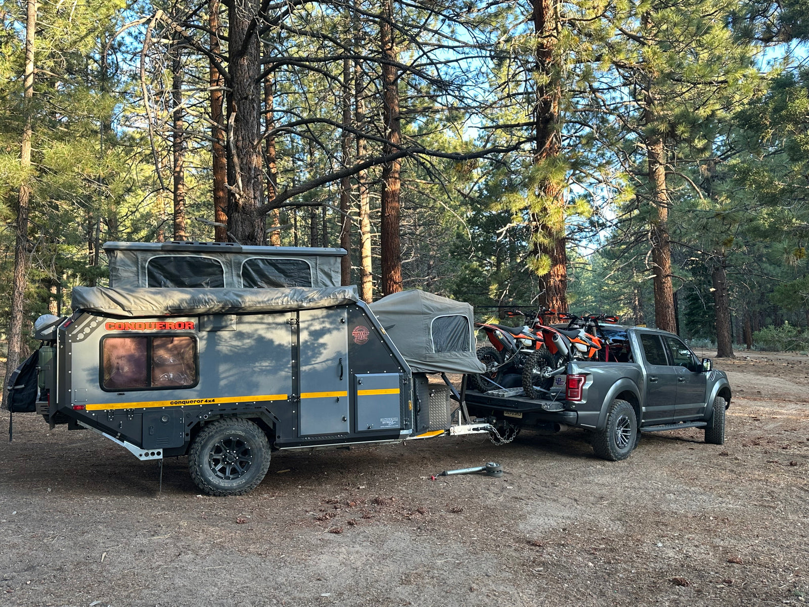 Dark gray off-road camper trailer, similar to an RV rental, hitched to a truck with dirt bikes, parked in a pine forest.