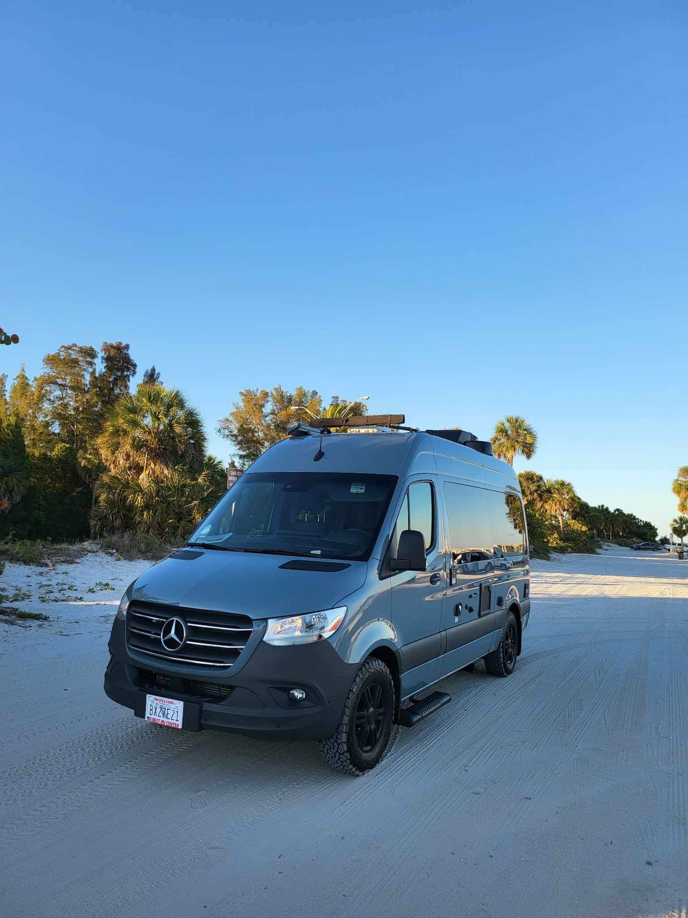 Gray Mercedes Sprinter RV rental on a sandy beach road among palm trees under a clear blue sky.