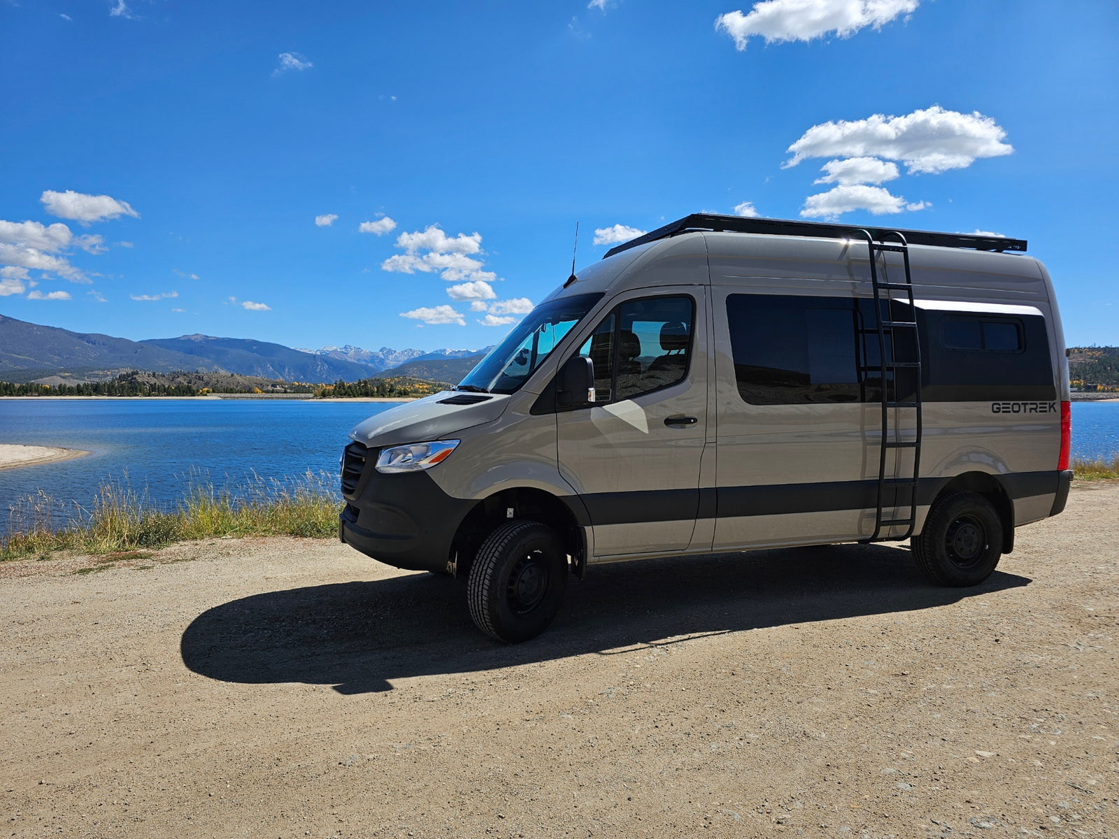 A light tan rv rental van, equipped for adventure, parked on a dirt road beside a blue lake with scenic mountains.