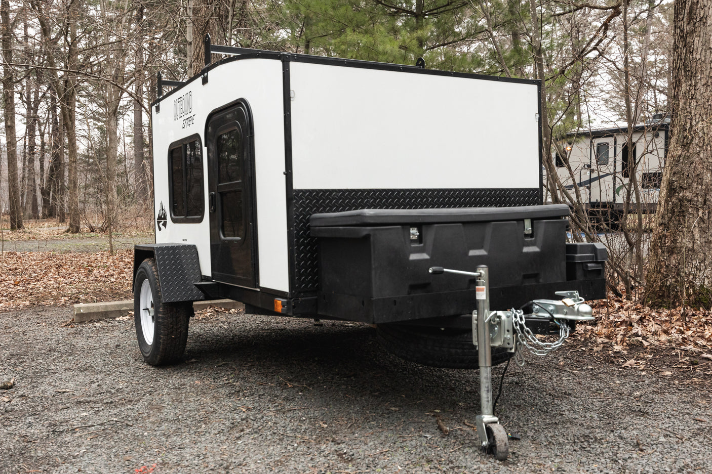This compact white and black "Outbound Extreme" camper, ideal for an RV rental adventure, is parked on gravel at a wooded campsite.