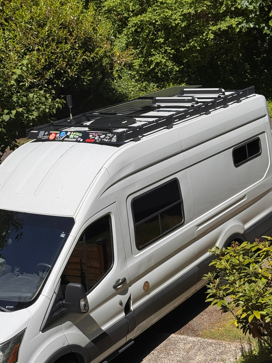A white camper van, ideal for RV rental, showcasing rooftop solar panels, a sturdy roof rack, and unique decals.