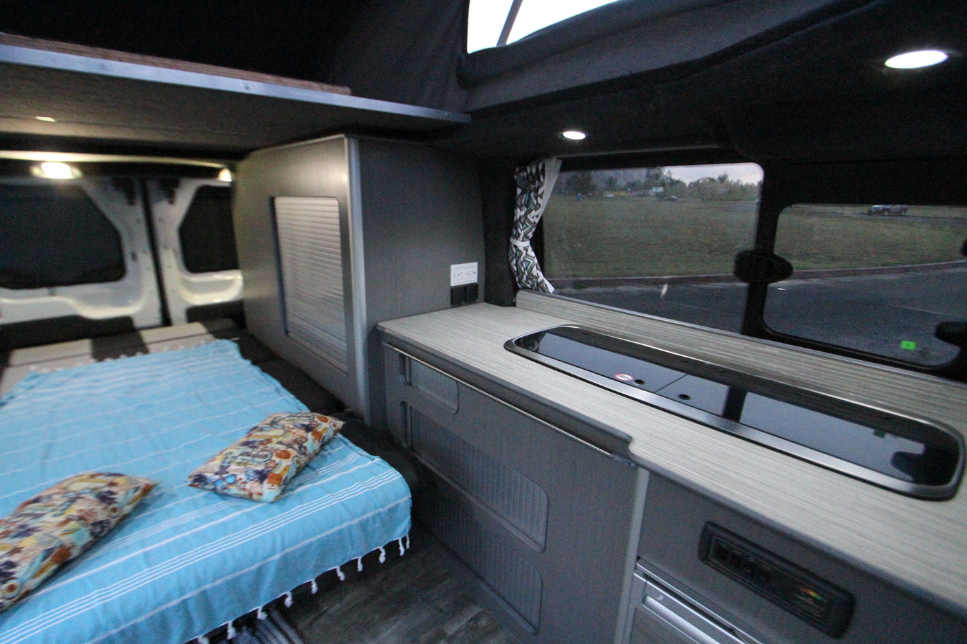 Explore this RV rental's modern campervan interior, featuring a cozy bed, sleek kitchen area, and scenic window.
