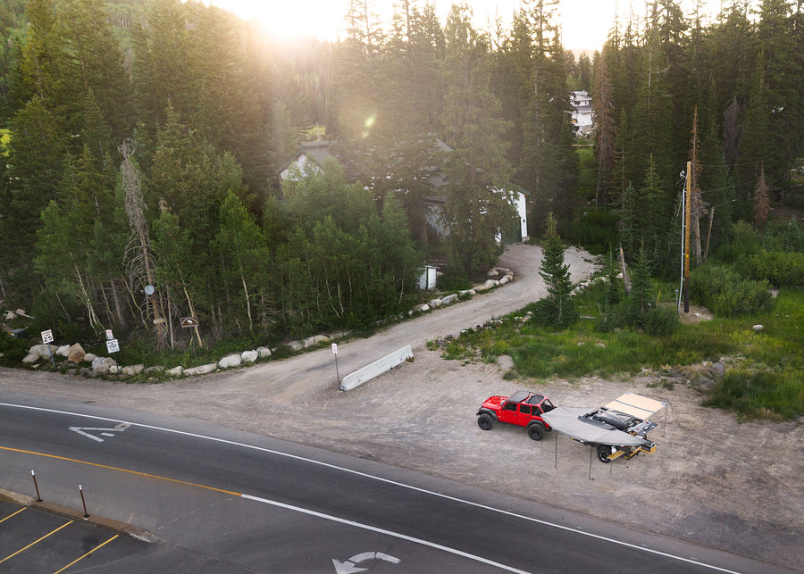 Red Jeep & compact camping trailer with deployed awnings by a scenic road in a forest. A unique RV rental for wilderness trips.