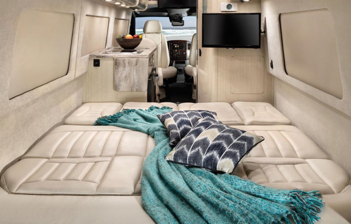 Enjoy this spacious RV rental camper van with a cozy bed, plush blanket, and a scenic beach view through the front.