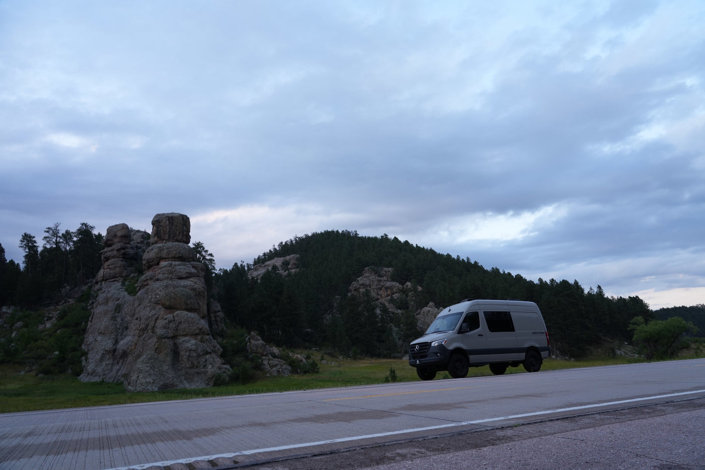A gray Mercedes Sprinter rv rental parked on a mountain road beside rock formations, pine trees, and cloudy skies.