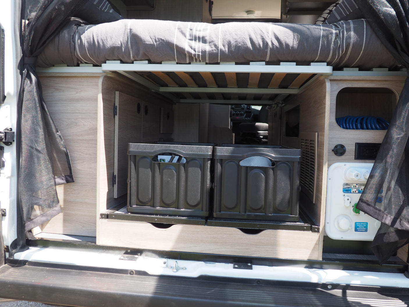 Rear view of an RV rental's organized living space, featuring a raised bed, sliding storage crates, and water hookups.
