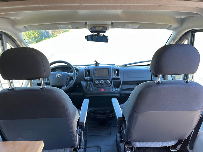 RV rental cockpit view from passenger area, showing front seats, dashboard with steering wheel, and road through windshield.