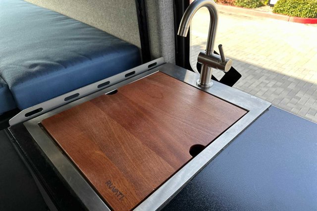 Inside an **rv rental**: a compact kitchen features a stainless steel faucet, sink with a Ruvati cutting board, and blue bed.
