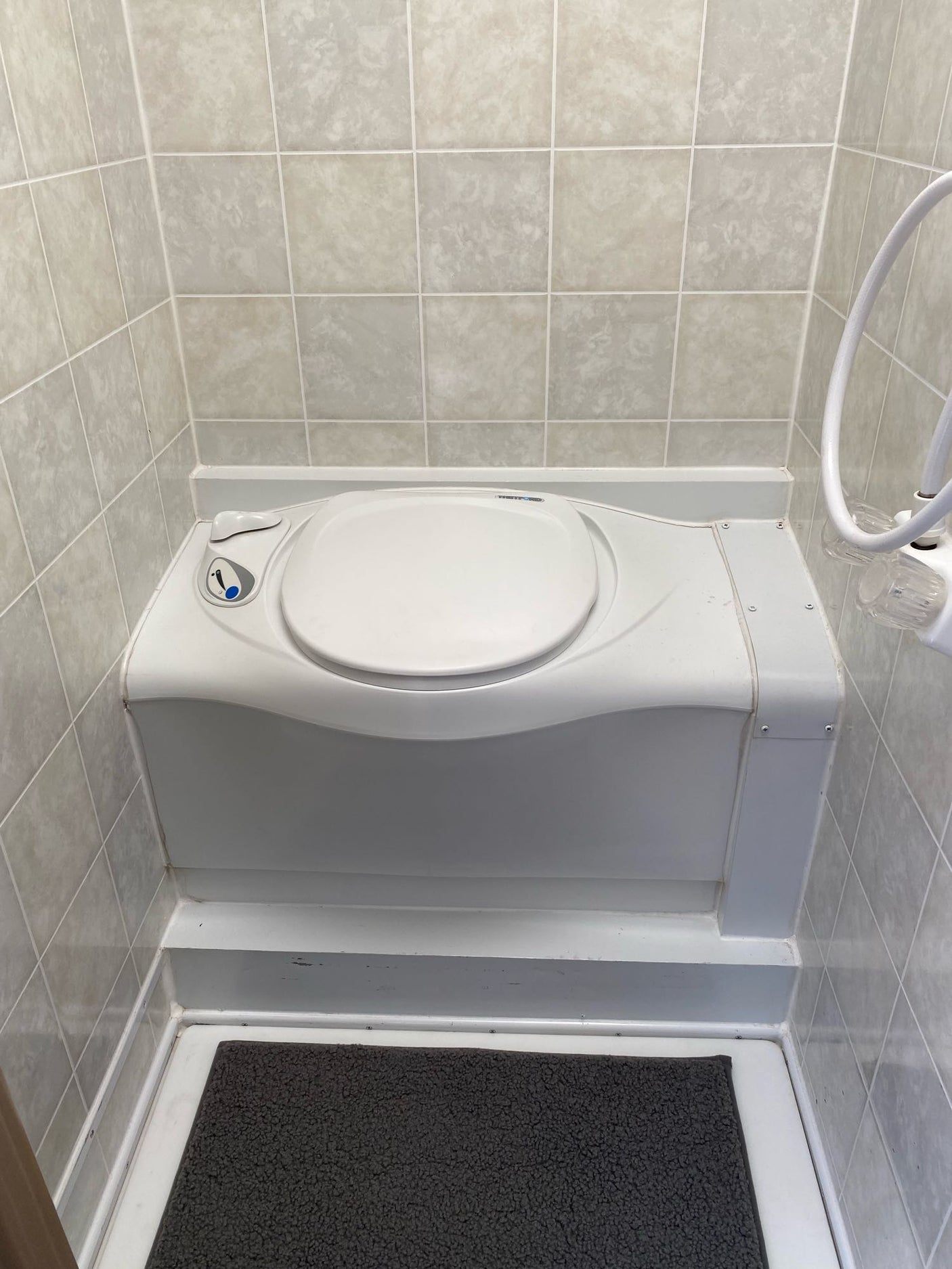 This RV rental's compact bathroom features a white toilet, tiled walls, a handheld shower, and a dark bath mat.
