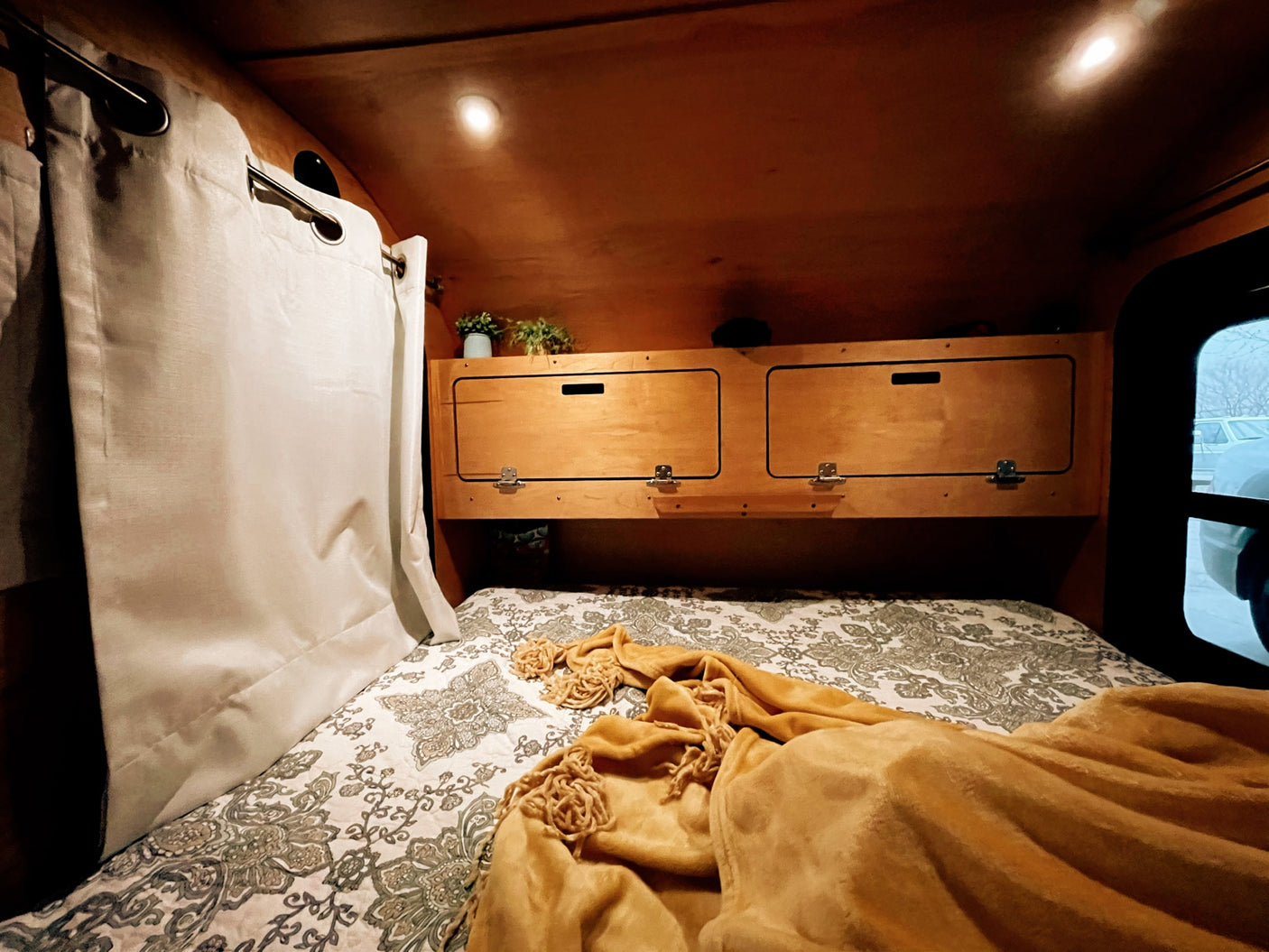 Cozy RV rental interior with a patterned bed, wood storage, bright lights, and window view, ideal for travel.
