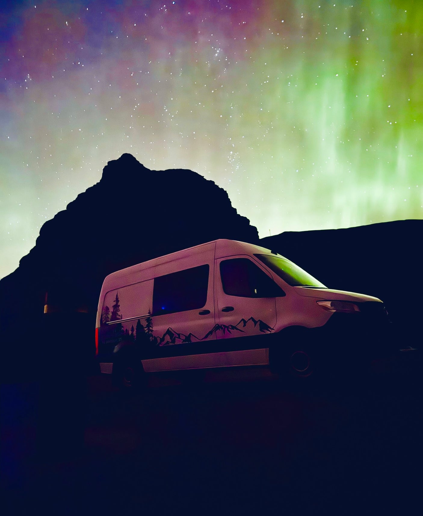 Green aurora & stars illuminate an RV rental camper van with mountain decals, parked before dark peaks.