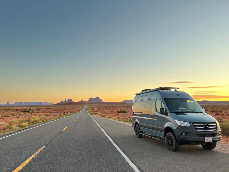 Scenic RV rental road trip: Grey Sprinter van on a desert highway with iconic mesas at a vibrant sunset.