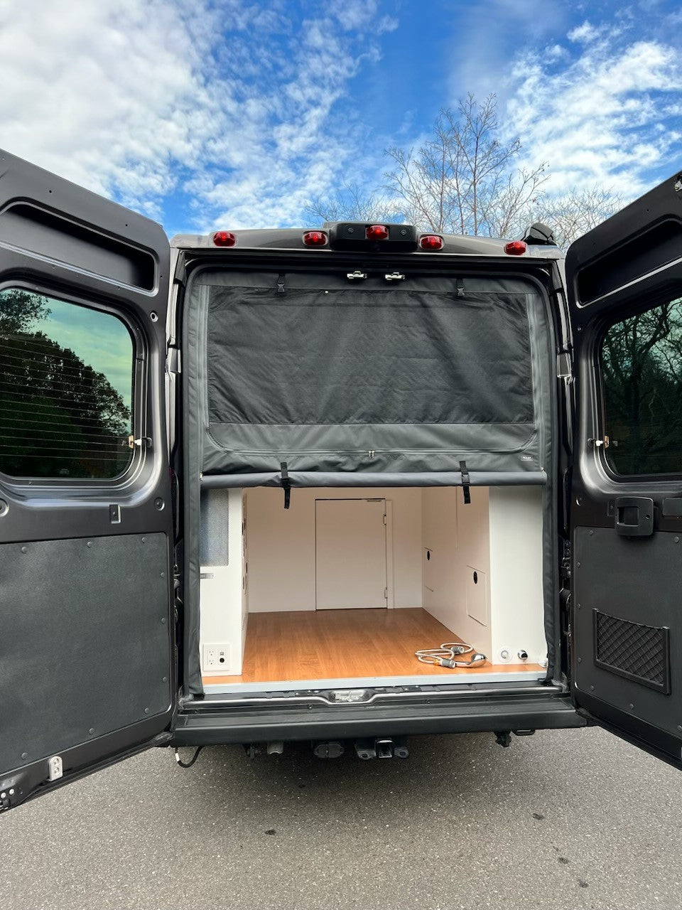 RV rental: Rear of a camper van with open doors, revealing a custom interior featuring wood flooring, white cabinets, and a black shade.