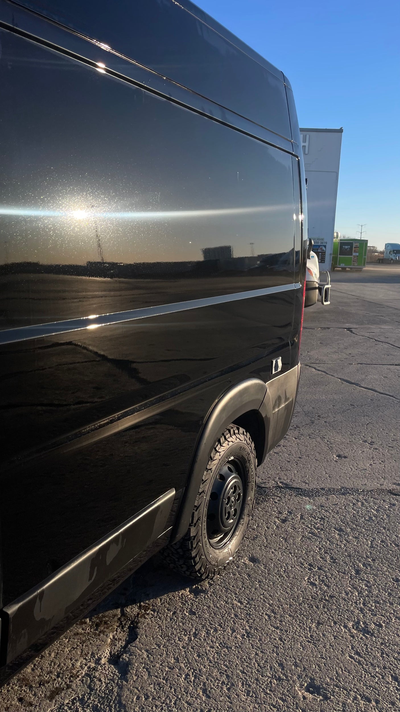 Side view of a black van with rugged tires, reflecting bright sunlight and showing specks on its surface. Great for an RV rental.