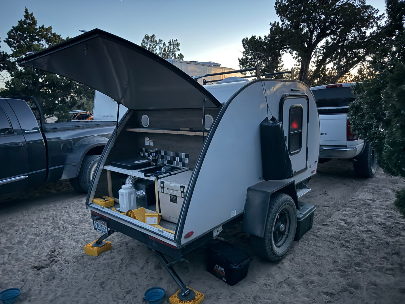 Modern teardrop rv rental camper with an open outdoor kitchen setup, parked on sand alongside a large truck.