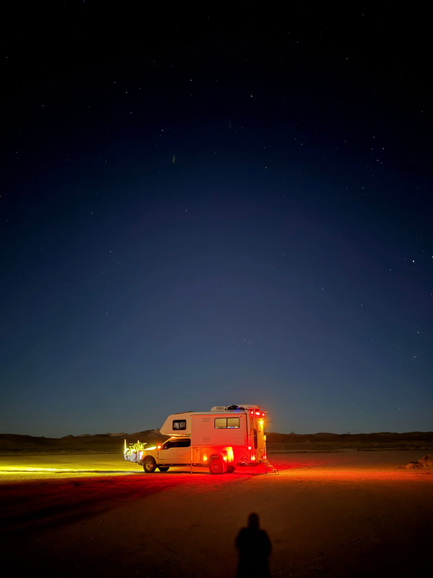 Starry night over an rv rental. White truck camper lights up desert ground; person's shadow in foreground. Epic boondocking!