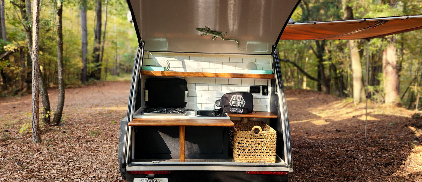 RV rental: Back of a teardrop camper showing a compact kitchen with stove, sink, and wood shelves, set in a peaceful forest.