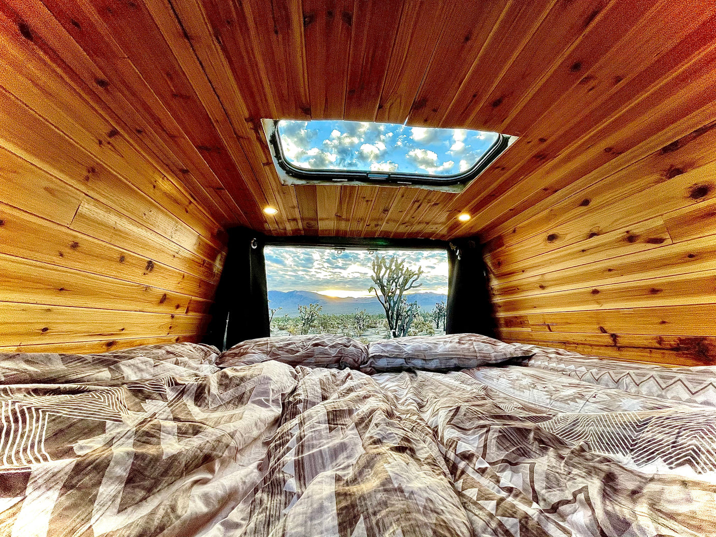 Cozy RV rental interior with bed and wood paneling, offering stunning desert sunset views and a sky through the roof.