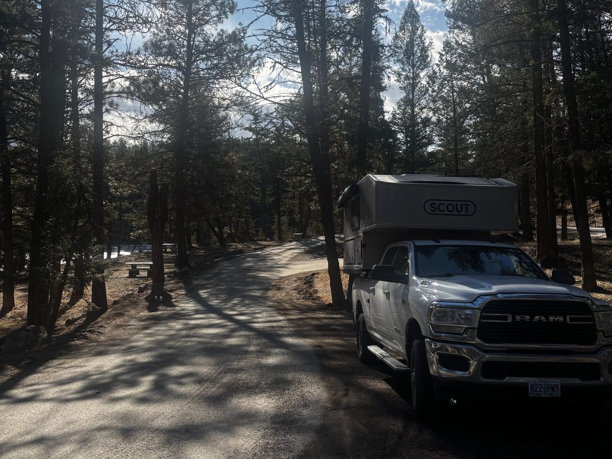 attached to a Ram pickup truck in a lush forest campsite. Ideal RV rental for outdoor adventures.|A white Ram truck with a Scout truck camper — photo 3