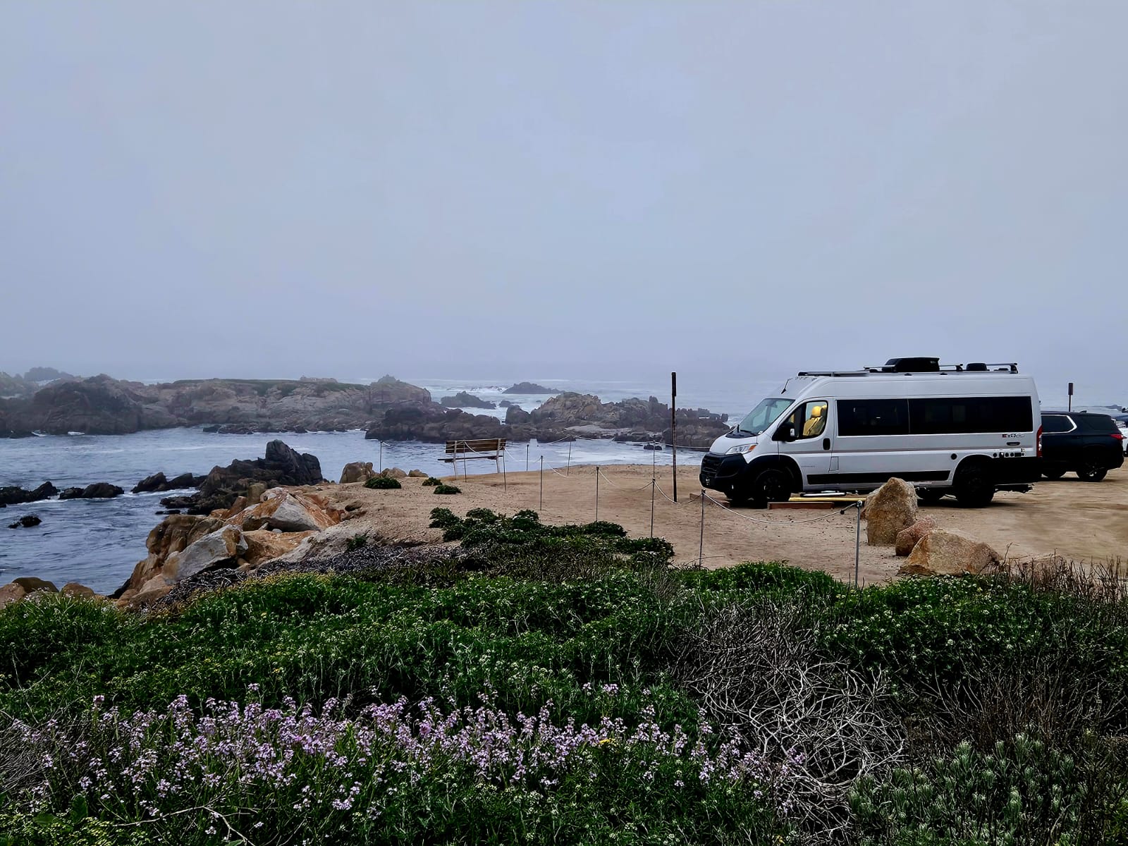 A white RV rental van and SUV parked on a misty, rocky coast overlooking the ocean with waves and a bench.
